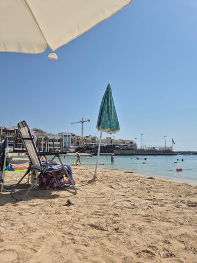 The image depicts a sunny day at a beach in Birżebbuġa, Malta. In the foreground, two beach chairs are set up on the sand, one with a blue and white striped pattern, and the other partially obscured and covered with a beach towel featuring a flamingo pattern. A closed, teal-colored umbrella with white fringe stands to the right of the chairs. Beyond the chairs, the shallow, clear water of the sea stretches out, with several people swimming and enjoying the water. Orange buoys mark a designated swimming area. In the background, a coastal town with multi-story buildings lines the shore, with palm trees visible amongst the architecture. A construction crane is prominently visible in the distance, suggesting ongoing development in the area. The sky is a clear, bright blue.