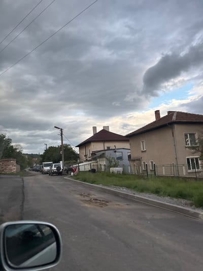 The image shows a street scene in Baylovo, Bulgaria. Buildings line the right side of the asphalt road. Overhead power lines are visible against a cloudy sky. A row of white utility trucks are parked along the roadside. A car's side mirror is in the bottom right corner of the frame.