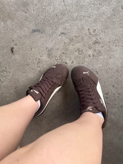 An overhead, medium shot shows a person's legs and feet wearing brown Puma sneakers with white accents and white socks, resting on a concrete surface. The concrete appears weathered and uneven, with a small, dark, cylindrical object lying on it to the left of the frame. The shot is taken from a high angle, looking down at the subject's legs and feet. The location is stated to be in Shulhynka, Ukraine.