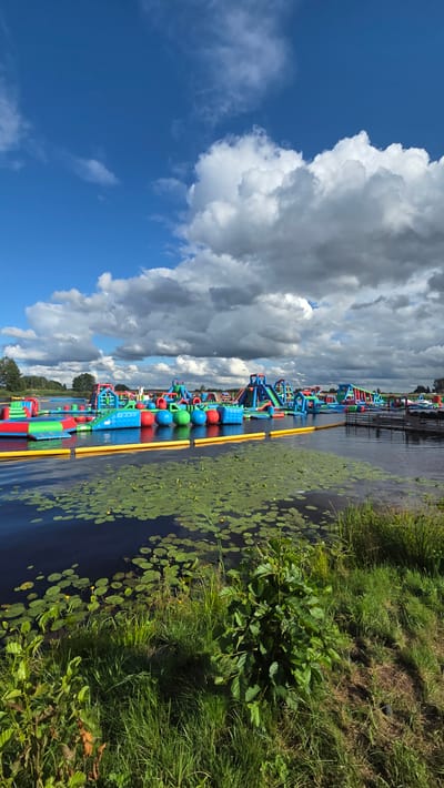 This is a medium shot, vertical, eye-level, natural light photograph of an inflatable water park situated on a lake in Jaunmārupe, Latvia. The park is composed of numerous brightly colored inflatable structures, including slides, climbing walls, and obstacle courses, all interconnected and floating on the water. Yellow inflatable buoys delineate a perimeter around a section of the park. The water is dark blue and partially covered with green lily pads. The foreground features a grassy embankment with lush green vegetation, including tall grasses and a leafy shrub. In the background, trees and a clear blue sky with scattered white and gray clouds frame the scene. No people are visible.