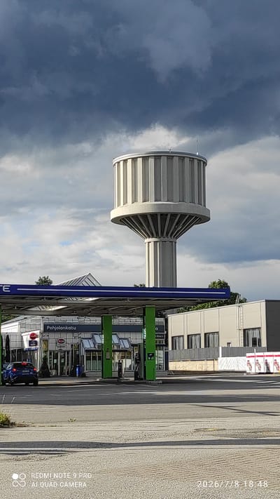 The image shows a water tower in Iisalmi, Finland, located at Pohjolankatu. The water tower is a tall, cylindrical structure with a fluted design, supported by a flared base and a single, thick column. In the foreground, a gas station canopy with green pillars and blue trim is visible, with a blue car parked to the left and a modern building with a glass facade to the right. The sky is dramatic with dark, heavy clouds. The overall scene suggests an urban or suburban setting.