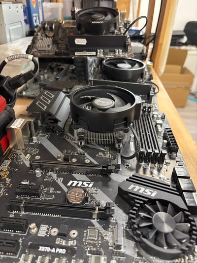 This is a close-up, low-angle shot of several computer motherboards, predominantly MSI brand, resting on a wooden surface. The motherboards are stacked or laid out horizontally, with some partially obscured by others. Visible components include CPU coolers with AMD branding, RAM slots, PCI-E slots, and various chips and capacitors. A power cable with a white label is also visible on the left side. The background shows shelves with boxes and what appears to be a server chassis. The lighting is natural, coming from the right side, casting shadows across the components. The scene is indoors, likely in a workshop or storage area. There is no indication of a specific location in Mülheim an der Ruhr, Germany.