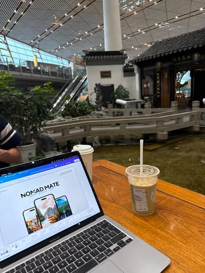 The image shows a laptop displaying a presentation titled "NOMAD MATE," a drink in a Starbucks cup with a straw, and another coffee cup on a wooden table. The laptop is a MacBook Air. The background reveals a building with traditional Chinese architecture and greenery, with a modern glass structure visible on the left. The image was taken at the Shunyi District, China.