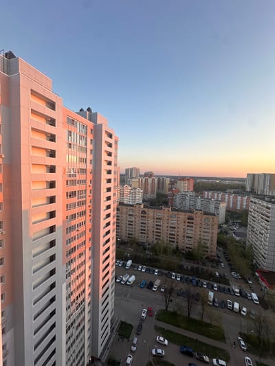 The image captures an elevated view of a city at either sunrise or sunset, likely in Gavrilkovo, Russia, given the prompt. The foreground is dominated by a tall, modern building with a pattern of rectangular windows and protruding architectural elements that create deep shadow lines, colored by the warm, horizontal light. Beyond the building, the cityscape unfolds, showing a mix of high-rise apartment complexes and lower-rise residential structures. Streets are visible with parked cars lining the sides. The sky transitions from a clear, pale blue to a gradient of orange and yellow near the horizon, indicating either a sunrise or sunset condition. The angle of the photograph suggests it was taken from a high vantage point, possibly from within another tall building or an elevated location. There are no discernible people visible in the frame. The overall impression is an urban landscape bathed in the soft light of dawn or dusk.
