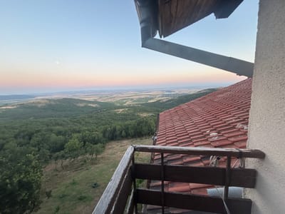 An outdoor shot captures a view from a balcony overlooking a vast, hilly landscape. The sky displays a gradient of soft colors, transitioning from a pale blue at the top to pink and orange hues near the horizon, with a faint sliver of the moon visible in the upper left. The foreground is dominated by a rustic wooden railing, showing signs of wear and tear. Beyond the railing, a red-tiled roof of a building extends towards the right side of the frame. The landscape unfolds with a dense green forest covering the hills, interspersed with patches of dry grass and some scattered trees. In the distance, the land flattens out, revealing agricultural fields and what appears to be a small settlement. The overall impression is one of a tranquil, rural setting, possibly during sunset or sunrise.