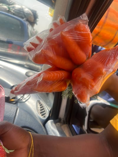 The image depicts an interior view from a vehicle in the city of Hoss, Nigeria. In the foreground, multiple small, unpeeled orange carrots are visible, each individually packaged in a transparent plastic bag and clustered together. A dark-skinned hand on the lower left holds a portion of a red fruit, likely a strawberry. A dark-skinned arm, partially obscured on the right, is clad in a yellow fabric. The vehicle's light grey dashboard, featuring a circular air vent, is visible in the mid-ground. Through the vehicle's window, a light blue sedan with bundles piled on its roof is discernible in the background, indicating an outdoor urban or market environment.
