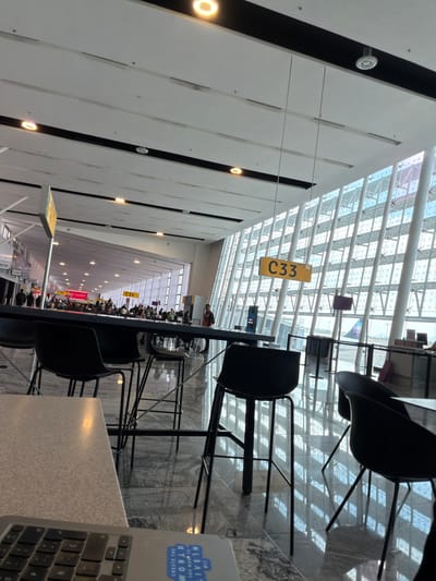 This image shows the interior of an airport terminal, possibly in Santa Cruz del Valle, Mexico, although location cannot be confirmed. In the foreground, there's a workspace with a table and black chairs. A laptop is partially visible on the right side of the frame. 

Overhead, a C33 sign is suspended, and further in the distance there is a C34 sign. Beyond the seating and working area, the terminal extends into the distance, filled with people waiting. The architecture features a high, geometric ceiling made of glass and metal, allowing natural light to enter.
