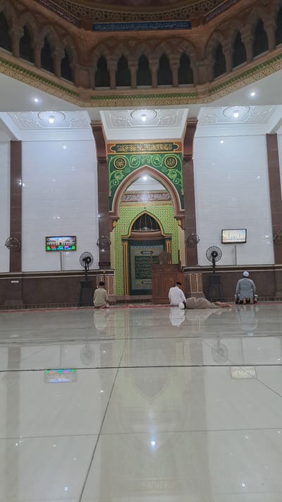 In the city of Lhokawe Teungoh, Indonesia, the image captures the interior of a mosque. Three men are present. One man, dressed in a light-colored shirt and black cap, is kneeling on a prayer mat, facing away from the camera. Another man, wearing a white shirt and black cap, is also kneeling on a prayer mat, facing away from the camera, with his head bowed in prayer. A third man, in a grey shirt and white cap, is kneeling on a prayer mat with his head bowed. The mosque features ornate architectural elements, including arched doorways with intricate carvings and patterned wall coverings. A wooden pulpit stands near the center of the room, and two large fans are visible on either side of the prayer area. A screen displaying text and an image is mounted on the wall to the left. The polished floor reflects the mosque's interior, creating a symmetrical visual effect.