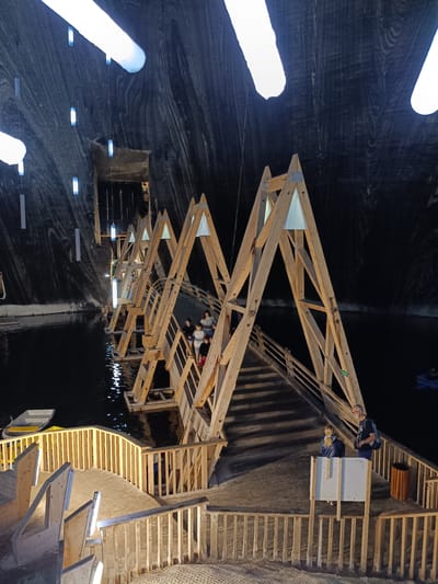 The image depicts an underground salt mine in Turda, Romania, transformed into a tourist attraction. The scene is dominated by the rough, striated walls of the salt cavern, illuminated by large, elongated light fixtures embedded in the ceiling and by smaller, rectangular lights hanging from wires. A dark, calm body of water fills the cavern floor, reflecting the lights and the wooden structure that spans it.
A prominent wooden bridge, constructed with a series of A-frame supports, crosses the water. The bridge is equipped with wooden railings and stairs, and several people are seen walking on it or standing at its edges. Some are looking out at the cavern, while others appear to be conversing. In the foreground, a wooden walkway with railings leads down to the water's edge, with several benches also visible. A small, white rowboat is moored near the walkway.
The lighting suggests it is likely nighttime or the cavern is artificially lit, as there is no natural daylight visible. The atmosphere appears serene and perhaps a bit awe-inspiring, given the unusual and grand setting. The overall impression is of a unique tourist destination that harmoniously blends natural geological formations with man-made structures and recreational elements. No discernible text is visible in the image.