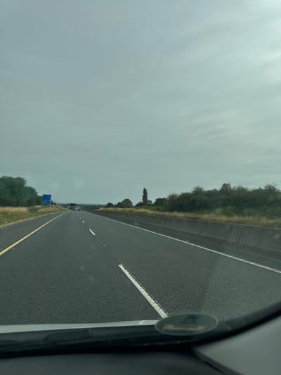 The image shows a view from inside a vehicle driving on a highway in Glaunta, Ireland. The sky is overcast and gray. The highway is paved with asphalt and has clear lane markings. There is a concrete barrier on the right side of the road and a grassy verge with trees on the left. A blue highway sign is visible on the left side of the road in the distance. There are a few other vehicles on the highway ahead. The lower part of the image shows the dashboard of the vehicle.