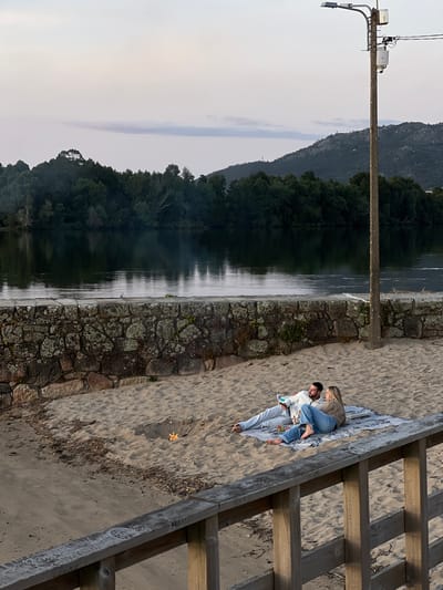 A landscape-oriented photograph displays a scene at Gondarém, Portugal. The image features a couple reclining on a blanket on a sandy surface next to a stone wall and adjacent to a body of water, most likely the Minho River.
On the left side of the image, a utility pole stands near the water’s edge. It has visible hardware and wires. A large expanse of water fills the mid-left, reflecting the dark green trees lining the opposite shore. Further in the background, a mountain range is partially obscured by a grey sky.
To the right, a stone wall divides the water from a sandy area. A couple is reclining on a light-colored blanket on the sand. The man has dark hair and a beard, wearing jeans and a white jacket. The woman, with blonde hair, is wearing jeans and a brown jacket, with her back to the camera.
To the right of the couple, there is a wooden stairway leading to the beach. A few small, sun-colored, objects are present on the sand near the stairway.