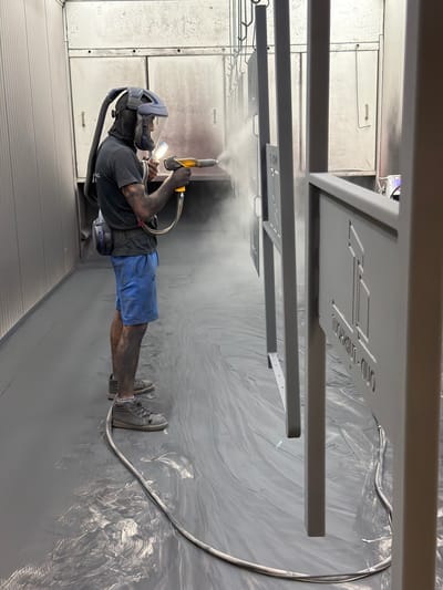A man wearing a protective helmet and mask is powder coating a metal sign in a dimly lit, industrial setting. The man is dressed in a dark t-shirt and blue shorts, with visible tattoos on his arms. He holds a spray gun in his right hand, emitting a cloud of grey powder onto the suspended sign. The sign appears to have cut-out lettering, and some text is partially visible, reading "DIAMOND - D..." and "ING". The floor of the setting is covered with a thin, crinkled material, likely a protective sheet, which is also dusted with the grey powder. Metal racks and shelving units are visible in the background, along with electrical conduits and cables. The overall atmosphere is one of focused industrial work. The lighting suggests an indoor environment, possibly a workshop or factory, with artificial lighting illuminating the scene. There are no clear indicators of the time of day or weather conditions. The location is identified as Mol, Belgium.