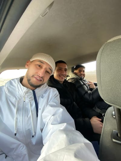 A photograph taken inside a vehicle depicts three adult males. The individual in the foreground, positioned to the left, wears a light-colored beanie and a white hooded jacket, appearing to hold the camera. He has a short beard and is smiling directly at the lens. Behind him, a second male with short dark hair and a light beard wears a dark puffer jacket and smiles towards the camera. Further to the right and rear, a third male, also with dark hair and a short beard, wears a dark puffer jacket over a light-colored hoodie; he is smiling and displays a peace sign with his right hand. The vehicle's interior shows a light-colored ceiling with a grab handle and a gray, textured fabric headrest in the bottom right. Through the vehicle's windows, a flat, light-colored field is visible under an overcast sky, suggesting an open, rural-like outdoor environment. The specific location within La Wantzenau, France, is not visually discernible from the image content.