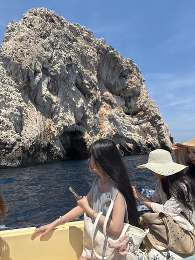 In the city of Komiža, Croatia, on a boat trip near a rocky cliff face with a cave opening, three women are captured in a medium shot. The woman on the left, facing away from the camera, has long, dark hair and wears a light blue dress, a silver necklace, and a heart-shaped charm bracelet. Her left hand rests on the yellow boat railing, adorned with a silver ring, while her right hand holds a clear phone case. Beside her, a woman in the center wears a wide-brimmed straw hat and a white lace top, with a tan suede purse at her side. Partially visible on the far right, another woman is seen wearing a straw hat. The boat, with its yellow railing, is on a dark blue body of water under a bright blue sky.