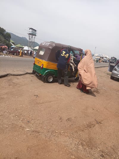 In Tilde, Nigeria, a dusty, unpaved road features a collision between a yellow and green three-wheeled auto rickshaw and the road's edge, causing the rickshaw to tilt precariously. A man in a dark blue shirt with a gold "34" on the back stands by the open door of the rickshaw, appearing to help load an animal, possibly a goat, into the vehicle. A woman in a beige hijab walks away from the rickshaw, her head and body covered by the garment. In the background, a water tower with multiple tanks stands tall, and other vehicles, including a silver car and several other auto rickshaws, are visible on a paved road to the right, suggesting a market or transportation hub. The sky is overcast.