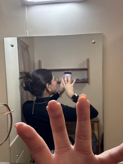 A mirrored image shows a person taking a selfie. The person is wearing glasses and a black top. A hand is held up towards the viewer. A window, bed and coat rack are visible in the background through the mirror. A pair of glasses is seen at the bottom of the mirror.