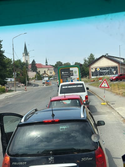 The image depicts a street scene in Dětmarovice, Czechia, likely during daytime with clear skies. The view is from inside a vehicle looking forward. Traffic is stopped or moving slowly, forming a queue.
In the foreground, the rear of a dark-colored Peugeot 307 SW is visible, with its driver's side rear door open. The car's tailgate and rear lights are prominent.
Further down the road, a red car is stopped, followed by a white van with blue flashing lights and text on the rear that reads "SMLUVNÍ ZDRAVOTNICKÉ ZAŘÍZENÍ". This suggests it is an ambulance or a medical transport vehicle. Next to the van is a green truck with advertisements.
The road appears to be undergoing construction or repair. There are construction barriers, cones, and visible rubble in some areas. A road roller is present on the right side of the road, suggesting ongoing work.
In the background, several buildings are visible. A prominent church with a tall spire and clock tower stands on the left side of the road. Another smaller church or steeple is visible further in the distance. On the right side of the road, there is a building with a sign that says "Pizza.cz" and depicts pizza slices. A red car is parked in front of this building.
Traffic signs are also visible, including a triangular sign with traffic light symbols and a diamond-shaped sign. Streetlights are present along the road. Trees and greenery line the sides of the road, indicating a semi-urban or suburban setting. The overall impression is a typical traffic situation with some roadworks in progress in a town with noticeable church architecture.