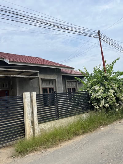 The image shows a house in Tanahterban, Indonesia. The house has a red tiled roof and gray concrete walls. It is surrounded by a black metal fence. In front of the fence, there is a large bush with white flowers. The sky is cloudy. A red utility pole with many wires runs across the top of the image. The road in front of the house is paved. There are no people visible in the image.