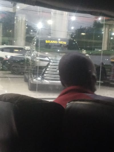The image displays an interior perspective looking out through a large glass window onto an exterior scene in Majengo ward, Kenya. In the foreground, the upper back and head of a seated individual are visible from behind. The person has dark hair and is wearing a red-collared garment beneath a darker outer layer. The exterior, indicative of a car dealership or showroom at night or dusk, features a dark-colored SUV or truck with a prominent chrome grille positioned centrally. Yellow text, "BRAND NEW," is discernible across the vehicle's upper front section. A white SUV is partially visible to the left. The scene is illuminated by multiple bright artificial lights, causing distinct starburst reflections on the glass. Trees and additional urban lights are visible in the background.
