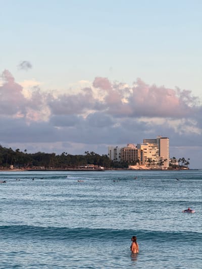The image depicts a coastal scene in Honolulu, United States of America. The foreground shows a stretch of blue water where several people are swimming and surfing. In the midground, a line of trees runs along the shore, with buildings visible behind them, including some taller structures that suggest a developed urban area. The sky, which occupies a significant portion of the image, is a mix of blue and clouds with hints of pink, indicating either sunrise or sunset. The photograph is presented in a rotated orientation.