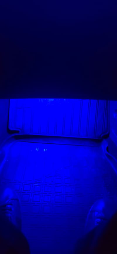 The image shows the interior of a vehicle, possibly a car, illuminated by blue LED lights. The floor mats appear to be black rubber with a textured pattern. There are hints of what might be feet or shoes in the lower corners, suggesting the perspective is from someone sitting in the vehicle. The upper portion of the image is dark, indicating the ceiling or upper interior of the vehicle. There are no discernible landmarks or external features that would allow for the identification of a specific location in the city of Enem, Russia.
