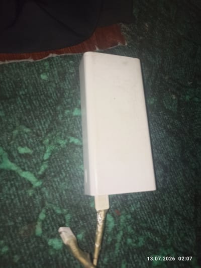 A white power bank is resting on a dark green textured surface. A cable is connected to the bottom of the power bank. In the background, a dark fabric is visible. A date and time stamp "13.07.2026 02:07" is present in the lower right corner of the image. The location is Chukurak, Tajikistan.