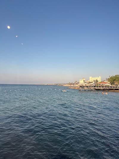 This image captures a picturesque scene of the Lapithos coastline in Northern Cyprus. The frame is divided into two distinct halves: the left side depicts a vast expanse of clear blue sky, gradually transitioning to a lighter hue near the horizon. Three luminous, circular objects, possibly balloons or orbs, are visible in the sky. The right side of the image is dominated by the shimmering, deep blue waters of the sea, with gentle waves creating a textured surface.

Along the shoreline, a charming coastal town unfolds. Buildings with white facades and red-tiled roofs are nestled amongst lush green trees. A pier extends into the water, dotted with people engaged in leisure activities. Several individuals are seen on paddleboards, enjoying the calm sea. A few boats are also present, further enhancing the sense of a vibrant seaside community.

The overall mood is serene and idyllic, suggesting a beautiful sunny day, likely late afternoon given the warm light and the position of the sun implied by the illumination of the orbs in the sky. The clarity of the sky and the tranquil state of the water indicate pleasant weather conditions. The presence of buildings, the pier, and people on watercraft clearly establish the location as a popular coastal destination. There is no visible text in the image.