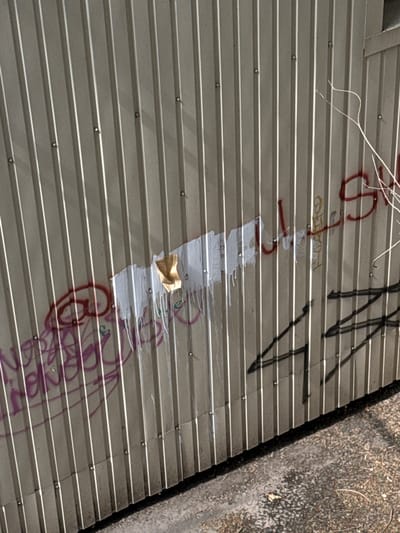 The image shows a close-up, eye-level, exterior shot of a corrugated metal wall covered in graffiti. The wall is predominantly gray with vertical ridges. In the center of the frame, a section of graffiti has been partially painted over with white paint that is dripping down the wall. A small piece of tan paper or tape is adhered to the white paint. Various graffiti tags in different colors, including pink, red, yellow, and black, are visible on the wall. To the right of the white paint, there are thin, white, vine-like objects or wires hanging down. The ground in the foreground is covered with gravel and dried leaves. The scene appears to be in an outdoor urban or industrial area. The location is identified as Zhovte, Ukraine.