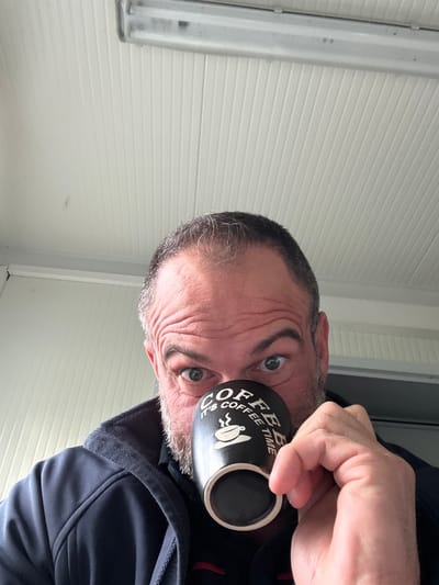 A medium shot depicts a fair-skinned man holding a black mug with white text that reads "Coffee Time" in front of his face. He has grey hair, blue eyes, and facial wrinkles, with a small amount of beard visible. He is wearing a dark blue hooded sweatshirt. The background is a white corrugated surface and a fluorescent light fixture. The location in Diekirch, Luxembourg cannot be identified from the visual data provided.