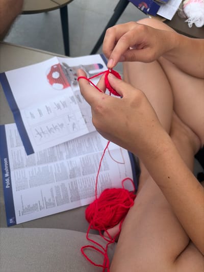 The image captures a close-up perspective of an individual engaged in crocheting within Grad Korčula, Croatia. The primary subject is a person's hands, actively working with bright red yarn and a silver crochet hook to form a small, circular red piece. An open instruction booklet lies flat on a light-colored surface beneath the hands; its visible title is "Poldi, Mushroom," and it includes an "EN" indicator for English. The booklet displays detailed textual instructions and diagrams, along with an illustration of the intended finished product, which appears to be a gray animal wearing a red and white spotted mushroom hat. A substantial ball of the same bright red yarn rests on the lap or adjacent surface. The individual's bare, tanned legs are partially visible in the lower right, and a hairy forearm belonging to another person is discernible in the upper left background, alongside blurred elements of an outdoor table and chair. The scene is brightly illuminated, suggesting an outdoor or well-lit indoor environment.