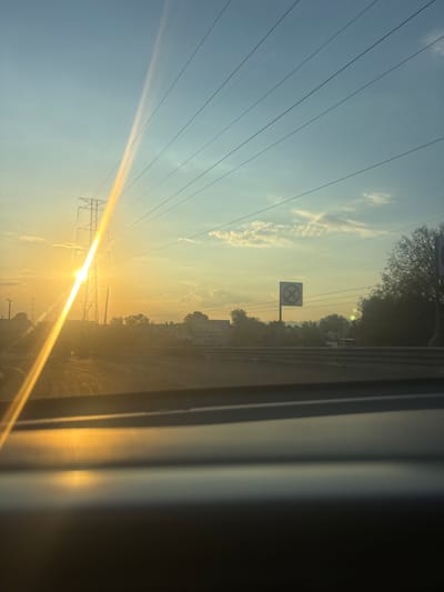 The image shows a view from inside a vehicle, possibly a car, during sunrise or sunset. The sky is light blue to orange, obscured by power lines and transmission towers. A railroad crossing sign with an "X" is visible. The location is likely Tultitlán, Mexico. The view is partially obstructed by the vehicle's interior.