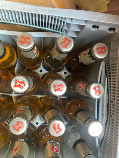 In Lütjenwestedt, Germany, a crate of Beck's beer bottles is depicted. The crate is white and plastic, with rows of slots designed to hold individual bottles. The bottles are made of brown glass and are filled with a golden-colored liquid, presumably beer. Each bottle is topped with a silver crown cap featuring a red shield with a white key symbol. The bottles are arranged vertically within the crate, with their tops facing upwards. Some of the bottles have labels around their necks, also displaying the Beck's logo. The lighting suggests the scene is indoors, possibly in a storage area or a retail space. The background consists of a wooden surface and a yellow cloth, adding context to the environment.