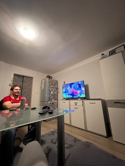 A man in a red jersey sits at a glass table in a room in Benningen, Germany. A television displays a cartoon. A refrigerator covered with magnets is in the background. The lighting is provided by a ceiling light. The room includes modern white cabinets.