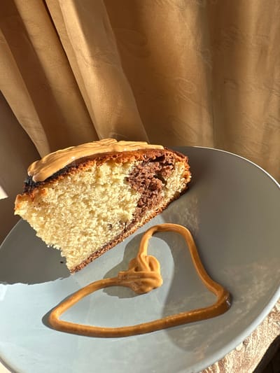 A slice of marbled cake, exhibiting a light and dark brown pattern, rests on a light blue-grey plate. The cake slice is topped with a light brown, viscous substance. An additional trail of this same light brown substance is dispensed onto the plate surface, forming an irregular, curled shape with a central knot, casting a distinct shadow. The scene is illuminated by strong, warm-toned light originating from the upper left, producing prominent shadows from the cake and the substance on the plate. The background features a draped, light brown or gold fabric, likely a curtain, exhibiting subtle folds and texture. No individuals are present. The documented location is Fowlis Easter, United Kingdom.