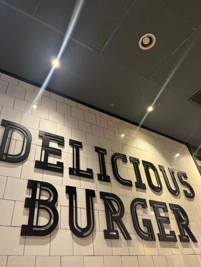 The image shows a vertically oriented, black-letter sign reading "DELICIOUS BURGER" mounted on a white-tiled wall. The sign is located inside a restaurant or food establishment, potentially in Kelapa Dua, Indonesia. The tiles are rectangular and appear to have dark grout lines. To the left of the sign is a dark ceiling with recessed lighting. There are visible reflections of light sources on the sign and ceiling.