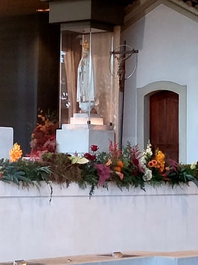 The image depicts an interior scene within the Sanctuary of Fátima in Fátima, Portugal. The central focus is a white, crowned statue of the Virgin Mary, consistent with Our Lady of Fátima, enclosed within a rectangular glass display case. This statue stands on a tiered white rectangular pedestal. To the immediate right of the glass enclosure, a wooden crucifix featuring the figure of Christ is mounted on a light-colored wall, framed by a circular metal ring. Below and to the right of the crucifix, a simple arched wooden door is set into the same wall. The foreground is dominated by a long, white rectangular structure, likely an altar or barrier, adorned with an extensive floral arrangement. This arrangement comprises various green foliage, red roses, white flowers, orange and yellow blooms, and reddish-purple leaves. Additional reddish-brown and orange foliage is visible behind the statue's pedestal. No individuals are present in the frame.