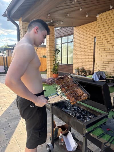 A young, shirtless man with dark hair and black cargo shorts stands beside a black barbecue grill outdoors. He is holding a metal grill basket filled with cooked meat over the grill, which contains hot coals. The man is facing the grill and looking down at the meat. The background shows a brick house with a wooden overhang, a paved patio, and some potted plants. String lights are visible hanging from the overhang. The location is identified as Dubovoye, Russia.