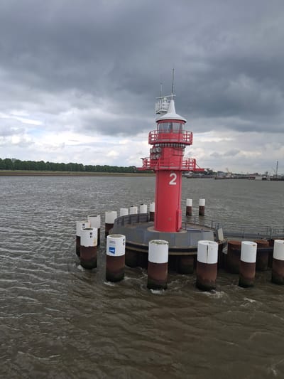The image shows a red lighthouse with the number "2" prominently displayed, situated in the Elbe River near Brunsbüttel, Germany. The lighthouse is mounted on a platform supported by several cylindrical pilings, many with white tops and identifying marks. The river is choppy with a muddy brown color. The sky is overcast with dark, heavy clouds. In the background, the shoreline is visible, with industrial structures and port facilities on the left, and a dense line of trees on the right. The overall impression is of a functional maritime structure in a working port environment on a cloudy day.