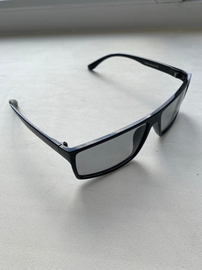 A pair of black-framed sunglasses with grey lenses rests on a textured white surface, possibly a windowsill or countertop, in an indoor setting. The sunglasses are positioned at an angle, with the left temple extending towards the bottom left of the frame and the right temple angled towards the upper right. A faint inscription is visible on the inner side of the right temple. The background is a blurred horizontal band of grey, suggesting a window or a wall. The lighting indicates a natural light source from above or the side. The scene is devoid of any people or discernible activity. The location is described as "Бескөл ауылдық округі, Kazakhstan," but this information does not correspond to visual elements within the provided image.