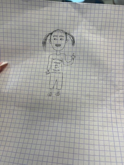 The image displays a hand-drawn cartoon figure rendered in dark ink on a sheet of white graph paper with blue grid lines, located in Ness Ziona, Israel. The paper exhibits minor creasing, primarily along its left edge. A partial human hand, featuring light-colored manicured fingernails, is visible in the lower-left quadrant, positioned as if holding or resting adjacent to the paper.

The drawing depicts a full-body humanoid character with a rounded head, two distinct pigtails, basic facial features including eyes gazing towards the figure's right, a simple nose, and an open mouth with visible teeth. The figure is clothed in a long-sleeved shirt bearing the inscription "NOT friendly please do NOT Touch". The figure's right arm is bent at the elbow, with the hand forming a gesture where the index finger points upwards. The left arm is also bent, with an open hand and splayed fingers. The lower body is clad in shorts or cropped trousers and shoes.