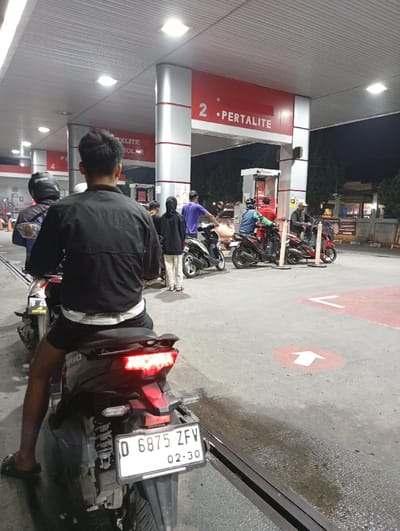 A queue of motorcycles waits at a gas station at night. The red and white station advertises "PERTALITE." A man in a black jacket sits on the first motorcycle, facing away from the camera. The bike's license plate reads "D 6875 ZFV." Other motorcycles are lined up behind, and people are milling around. An arrow on the ground indicates the direction of travel. The lighting is bright, and the scene appears to be in Indonesia, based on the license plate.