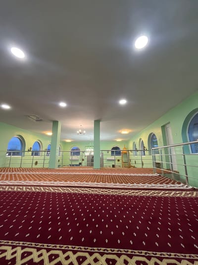The image displays the interior of a large, unoccupied hall, located in Sredkovets, Bulgaria. The perspective is from a low angle, looking across a raised platform towards a lower floor area. The foreground is covered by a dark red carpet featuring a repeating pattern of small white diamond shapes and a beige geometric border. Adjacent to this, further back, an orange-brown carpet with a similar pattern extends across the floor.

A silver-toned metal railing, composed of multiple horizontal bars and vertical supports, extends across the midground, separating the raised carpeted area from a lower floor section. Two prominent, square-sectioned pillars, painted a light mint green like the walls, support the ceiling in the central view.

The walls of the hall are painted a light mint green. The off-white ceiling is illuminated by multiple circular recessed lights and two chandeliers: one green-toned with multiple bulbs positioned centrally in the background, and another simpler, bronze-toned chandelier further to the left. Along the walls of the lower level, several arched window openings are visible, some revealing a dark exterior. In the distant background, a structure with intricate green and gold geometric patterns, consistent with a mihrab, is centrally positioned on the lower level. A white door with a window panel is present on the far right wall. No people or animals are observed within the scene.