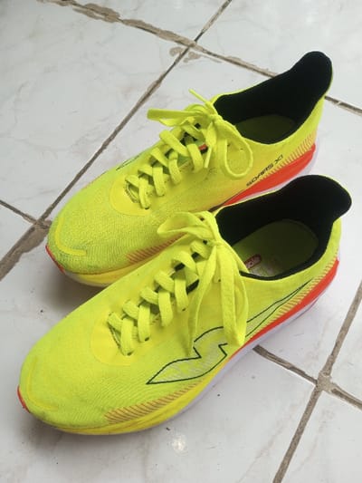 Two pairs of vibrant yellow running shoes sit on a tiled floor. Black lining is visible inside the shoes. The laces are also yellow. "SOARS XI" is printed on the side, near the heel. Orange accents highlight the heel and sides of the sole. The shoes appear modern and designed for speed.