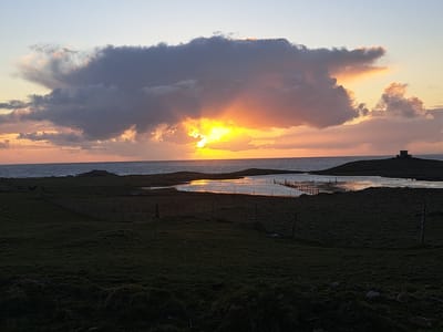 The image depicts a sunset over water in Balevullin, United Kingdom. A large, dark cloud obscures the sun, with rays of light breaking through. The sun reflects on the water's surface. The foreground shows land covered with green grass. Fences are visible along the water's edge, and a small tower-like structure is on the left. The sky is a mix of yellow, orange, and grey hues.