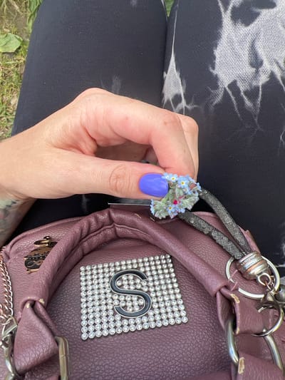 A person's hand with a purple-painted fingernail holds a small bouquet of blue forget-me-nots and green foliage. The hand is positioned over a mauve-colored purse adorned with a square of rhinestones that form a swirling design. The purse also has metal hardware, including a chain strap, rings, and charms. The person is wearing black leggings with a tie-dye pattern on the upper portion. The background shows green grass and a hint of dirt. The scene appears to be outdoors, possibly in a park or grassy area, under natural light. The person is likely showcasing the small floral arrangement or the purse.