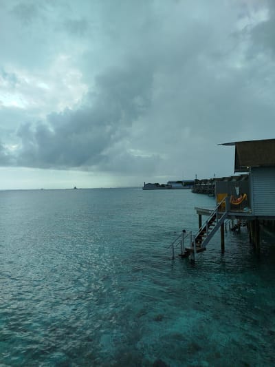 The image shows a view of the ocean at Gulhi Falhu, Maldives, under a cloudy sky. In the foreground, the turquoise water is clear enough to see the seabed. To the right, an overwater bungalow is partially visible, with a wooden deck and stairs leading down to the water. A person is relaxing in a hammock on the deck. In the distance, several other overwater bungalows are lined up along a jetty, and the coastline with buildings is visible on the horizon. A few boats, including a larger vessel that appears to be a ship or rig, are present on the sea in the far distance. The overall mood is serene and tropical, despite the overcast weather.