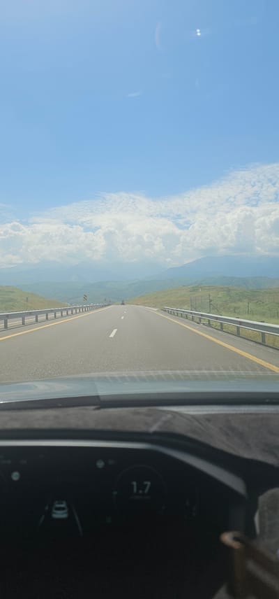A vertical, eye-level, point-of-view shot captures a car's perspective as it drives on a highway. The highway stretches into the distance, flanked by guardrails and lined with yellow markings. Rolling green hills and mountains form the backdrop, under a bright blue sky dotted with fluffy white clouds. In the foreground, the car's dashboard is visible, displaying a digital readout of "1.7" and a schematic of the vehicle itself. The scene suggests a drive through a scenic, likely rural, area. There is no visible indication of the specific location within Қырғауылды, Kazakhstan, other than it being a highway in a mountainous, green landscape.