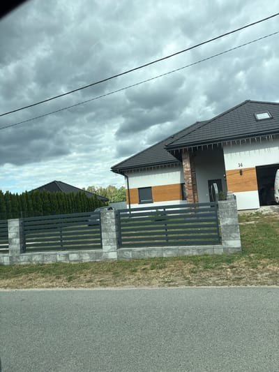 In the city of Mikorzyn, Poland, a modern single-family house with a dark-tiled roof and gray stone fence is pictured under a cloudy sky. The house features white and wood-paneled walls, with the house number "14" visible above the garage. A black car is parked in the open garage, and a fence with horizontal gray slats encloses the property. A row of tall green trees stands behind the fence, and a dark asphalt road is in the foreground. The sky is overcast with heavy gray clouds.