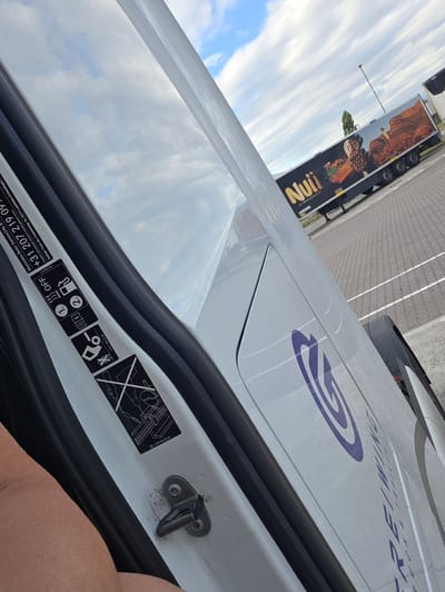 The image shows the interior of a white van, possibly a Mercedes-Benz, with the door open. A person's arm is visible at the top left, with light skin and some arm hair. The van's interior includes a black rubber seal along the door frame and a metal latch mechanism. Several stickers are affixed to the white surface inside the door. One sticker features a diagram of a vehicle with a person, and another shows icons for heat, an open book, and a fuel pump, with the text "OFF" and "1." and "2."
A prominent sticker displays "Mercedes-Benz Service24h" and a phone number "+31 207 219 09". Below this, there is smaller text indicating "Regulieren befonen kunnen in rekening gebracht worden" and "Daimler Truck AG, Fasanenweg 10, 70771 Leinfelden-Echterdingen". Another sticker shows a part number "A 001 584 72 38".
On the exterior of the van, a large purple "G" logo is visible, along with partially visible text that appears to be "GREI/WING TRUCKS".
To the right of the van, a truck trailer is parked, branded with the "Nuii" logo and an advertisement for "ICE CREAM ADVENTURE" featuring Australian landscape imagery. The truck is black with large graphics of ice cream and a desert scene.
The scene appears to be outdoors, with a cloudy sky visible in the reflection on the van's surface and in the background. The ground is paved with a patterned surface, suggesting a parking lot or service area. The overall impression is that this is a daytime shot, likely taken in Flörsheim, Germany, where the Mercedes-Benz truck division is located. The mood is neutral, focusing on details of the vehicle and its branding.