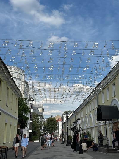 This is a street-level, vertical-format, eye-level shot looking down a pedestrianized street in Putyatino (Semivragovskaya S/A), Russia. The sky is blue with scattered white clouds. Strands of white, illuminated musical notes are strung across the street, decorating the scene. The street is lined with two-story buildings, predominantly yellow with arched window details on the right side. On the left, the building is also yellow with visible windows.
People are walking along the street. In the foreground on the left, two young women walk towards the viewer, one in a gray jacket and white shorts, the other in a blue shirt and white shorts. Further down the street, a group of people are walking in various directions. On the right side of the street, there is a black awning over an entrance, with a railing and stairs. A sculpture of a cello and a figure appears to be on the sidewalk near a lamppost. Several individuals are gathered near this sculpture and the building entrance. The street pavement is visible, and lampposts are present along the sidewalk. Trees are interspersed among the buildings.